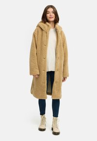 Long manteau en fausse fourrure beige avec capuche, fermeture à boutons et fentes sur les côtés. Porté sur un pull léger et un jean bleu, accompagné de bottes beiges à lacets.