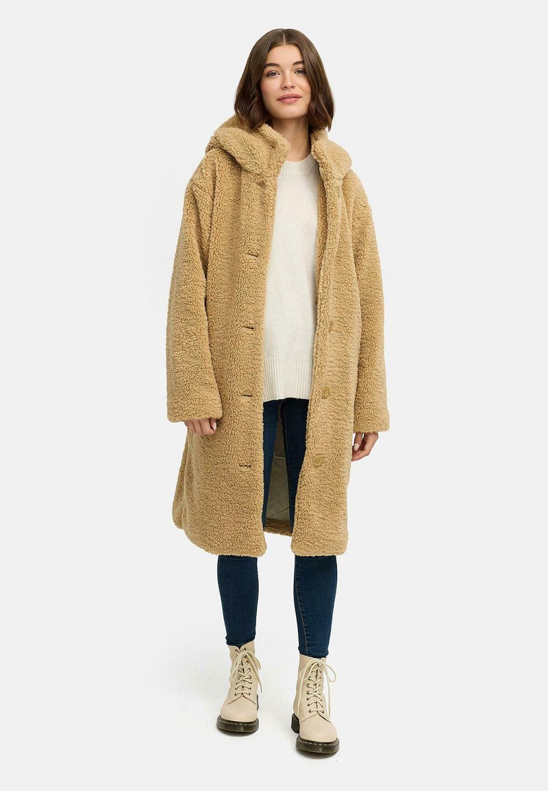 Long manteau en fausse fourrure beige avec capuche, fermeture à boutons et fentes sur les côtés. Porté sur un pull léger et un jean bleu, accompagné de bottes beiges à lacets.