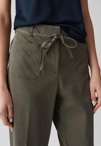 Olive green pants with a drawstring waist, two front pockets, and a smooth texture. Paired with a navy top, showing a relaxed fit.