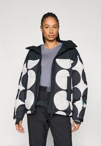 De donzen jas heeft een zwart-wit cirkelpatroon, ritssluiting aan de voorkant, verstelbare kap en zijzakken. Draagt over een grijs shirt en zwarte broek.