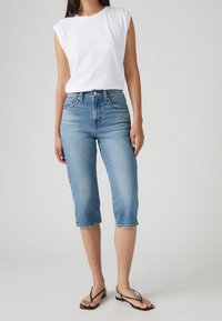 Světle modré džíny capri se slim fit designem, vysokým pasem, třepeným lemem a poutky na opasek, v kombinaci s bílým topem bez rukávů.