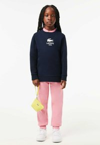 Sweatshirt navy avec logo Lacoste blanc ; col rose. Pantalons de jogging roses ; sac à main jaune texturé avec une petite poignée ; baskets blanches.