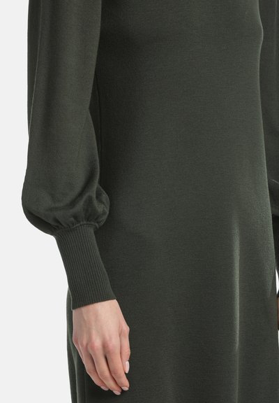 Dunkelgrünes Strickkleid mit gerippten Bündchen und puffigen Ärmeln. Nahaufnahme des Arms und der Stoffstruktur, die Weichheit und Dehnbarkeit hervorhebt.