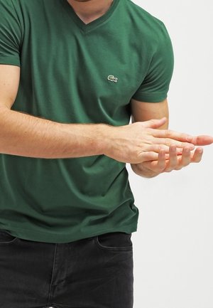 Mężczyzna w zielonej koszulce z dekoltem w kształcie litery V i czarnych spodniach, pocierający prawą dłoń lewą na jednolitym białym tle.