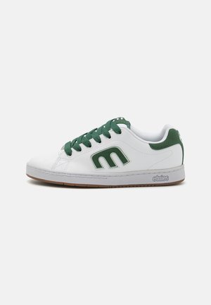 Sapatilhas brancas com realces verdes, apresentando um cabedal em couro liso, atacadores texturizados e sola de borracha. Logótipo da Etnies na lateral.