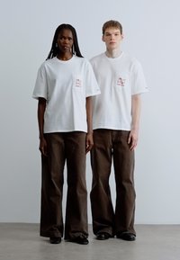 Camisetas blancas de algodón con bolsillos delanteros, combinadas con pantalones marrones sueltos. Diseño sencillo, sin patrones ni logotipos visibles. Se llevan zapatos planos.
