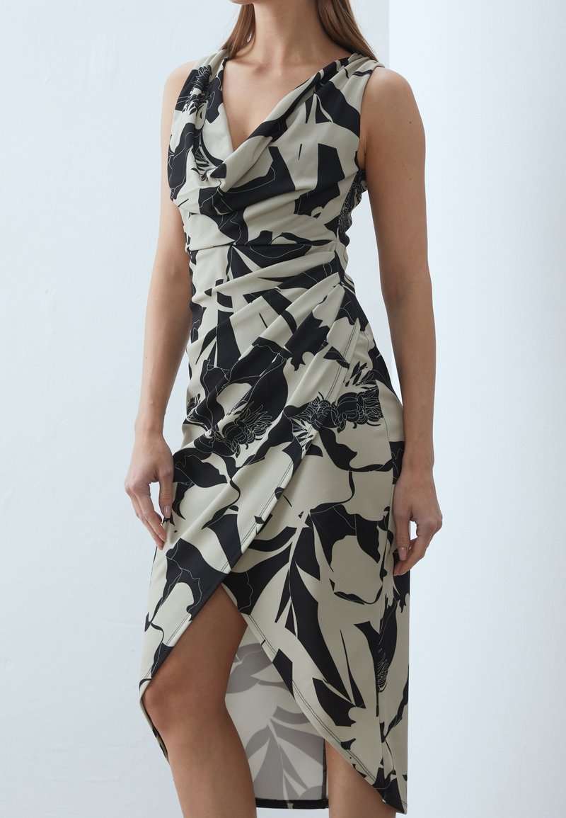 Robe beige sans manches avec imprimé floral noir, col bénitier et ourlet asymétrique dévoilant une jambe sur une femme debout.
