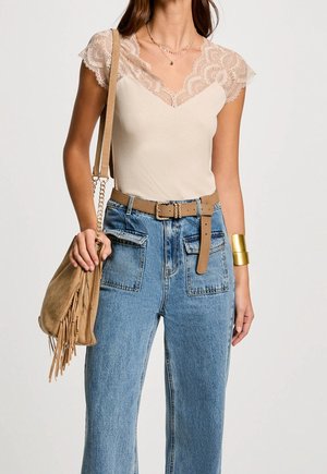 Kobieta w beżowej bluzce ozdobionej koronką, niebieskich dżinsach z wysokim stanem i kieszeniami naszywanymi, brązowym pasku, zamszowej torbie z frędzlami oraz złotej bransoletce na nadgarstku.
