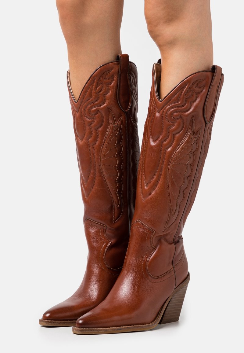 Botas de vaquero de cuero marrón con puntas puntiagudas, costuras intrincadas y cañas altas y anguladas. Diseño con tacón y detalles decorativos.