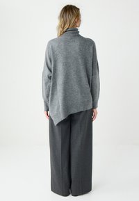 Szary, za duży sweter z golfem, miękka tekstura i skośny dół. W połączeniu z szerokimi, ciemnymi spodniami. Widok z tyłu.