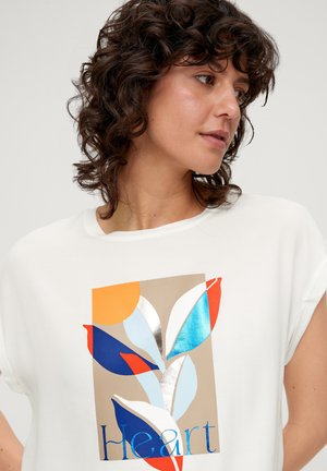 Chemise blanche à manches courtes avec un graphique coloré de feuilles en orange, bleu et argent métallique, portant le mot "Cœur" en texte bleu.