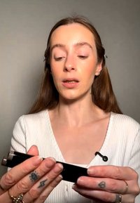 Nuori nainen, jolla on pitkät hiukset, pitää käsissään ja tutkii mustaa meikkituotetta, yllään valkoinen ribbineuloksinen toppi ja pieni mikrofoni kauluksessa.