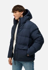 Giacca puffer blu navy con cappuccio, chiusura con zip e due tasche frontali. Presenta un design trapuntato e un dettaglio del logo sul lato sinistro del petto.