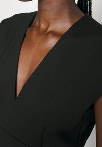 Musta mekko syvällä V-pääntiellä. Kangas näyttää sileältä ja jämäkältä, ja siinä on lyhyet hihat sekä minimaalinen yksityiskohtaisuus.