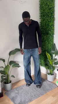 Man wearing black long-sleeve shirt and wide jeans stands on gray rug in a room with two potted plants and a green vertical wall panel.