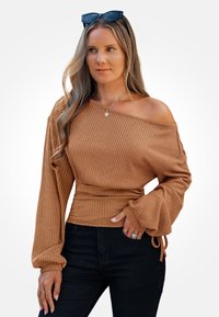 Pasiasty, brązowy top typu off-the-shoulder z długimi, rozszerzanymi rękawami i dopasowanym talią. Połączony z czarnymi spodniami i warstwowymi dodatkami biżuteryjnymi.