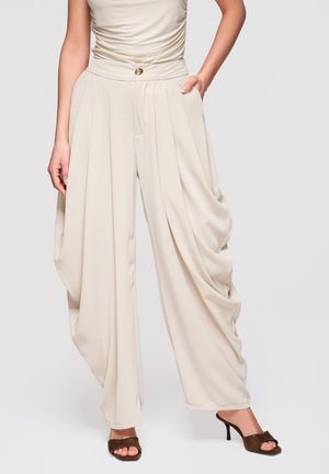 Kvinde iført beige bukser med høj talje og vide ben med draperede sidefolder og knappedetalje, kombineret med sorte åbne hæle.