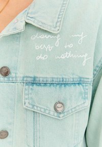 Jasnoniebieska kurtka dżinsowa z srebrnymi guzikami i białym odręcznym napisem: „robię co w mojej mocy, żeby nic nie robić” na górnej części klatki piersiowej.