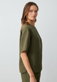 T-shirt in lino verde oliva con maniche corte arrotolate e scollo a barco. Il tessuto ha una texture sottile e una vestibilità rilassata.