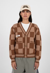 Femme portant un cardigan à carreaux beige et marron avec des boutons, un pantalon gris, une chemise blanche et une casquette beige portant l'inscription « Always Late Club ».