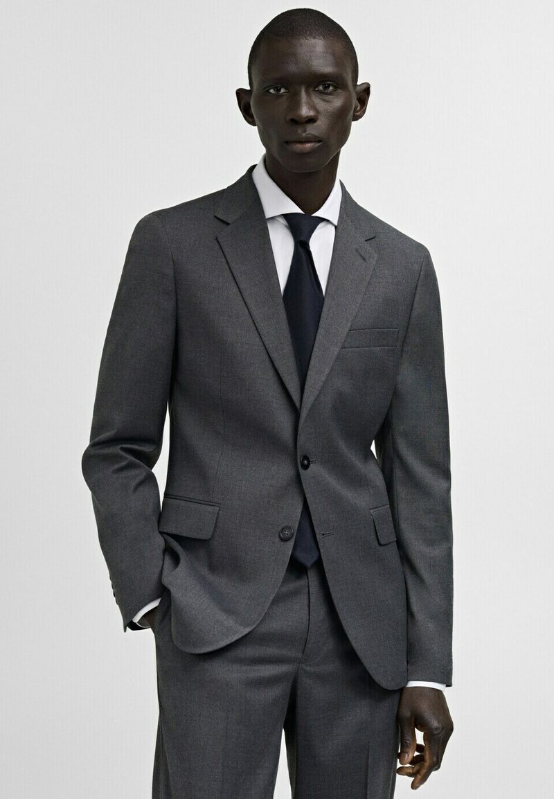 Homme portant un costume gris foncé sur mesure avec une chemise blanche et une cravate noire, debout devant un fond clair uni, les mains détendues.