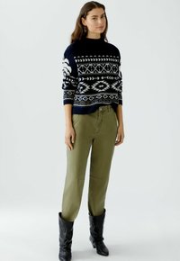 Donkerblauwe trui met witte geometrische patronen, gecombineerd met olijfgroene broek. Zwarte kniehoge laarzen maken de look compleet.