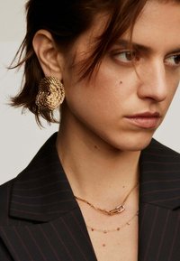Mujer con cabello corto castaño lleva un gran pendiente de oro texturizado, collares delicados de oro en capas y un blazer oscuro a rayas.