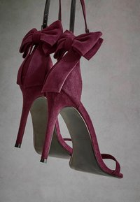 Sandales à talons hauts en velours bourgogne avec des accents de nœuds surdimensionnés, design à bout ouvert et fines brides de cheville. Talon et semelle noirs brillants.