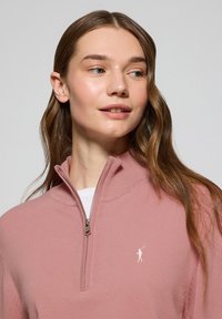 Pull léger rose avec un col zippé à mi-hauteur, texture côtelée et petit logo blanc. Doté de manches longues et d'une coupe décontractée.