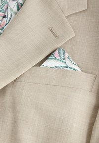 Manche de veste beige et poche poitrine avec un mouchoir de poche à motifs blanc, vert et rose.