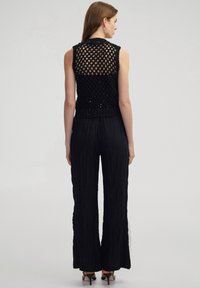 Donna in piedi di spalle, con indosso un top senza maniche in rete nera, pantaloni larghi neri e strutturati, sandali con tacco neri e orecchini a cerchio.