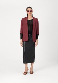 Blazer bordeaux avec bords festonnés, haut en dentelle noire transparente, jupe midi en cuir noir et sandales à talons brunes avec des accents décoratifs.