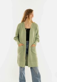 Vrouw met lang golvend haar die een lichtgroene gebreide cardigan en blauwe jeans draagt, glimlachend en naar de zijkant kijkend met haar handen in de zakken.