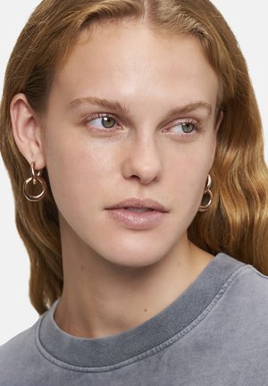 Goldfarbene Creolen mit zwei ineinandergreifenden Ringen. Das Model trägt einen grauen Sweatshirt, hat leicht welliges Haar und dezentes Make-up. Natürliches Licht.