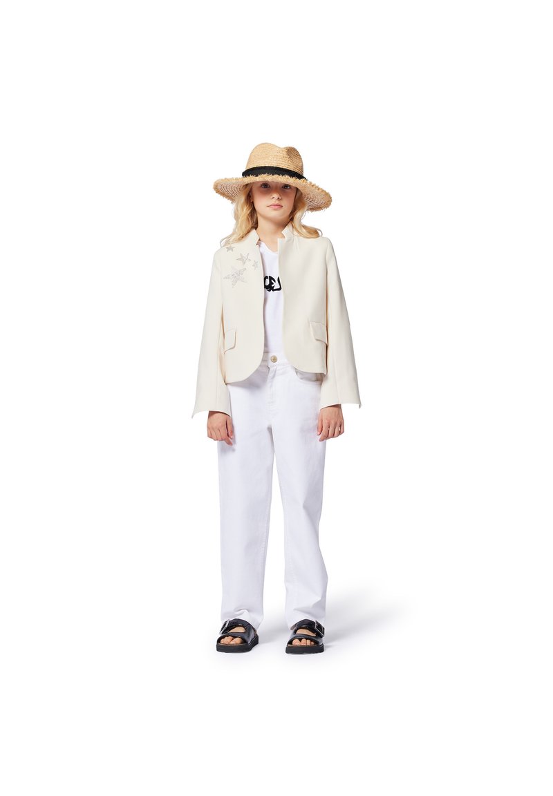 Witte blazer met zilveren steraccenten, wit T-shirt, losse witte broek en zwarte sandalen. Het model draagt een brede rieten hoed.