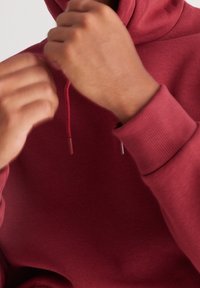 Burgundy huvtröja med hög krage, dragsko och ribbade ärmar. Tyget verkar mjukt och slätt med en något texturerad yta.