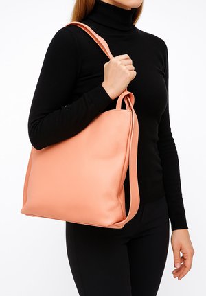 Donna con dolcevita nero che tiene una grande borsa a tracolla in pelle color pesca su una spalla, su sfondo bianco.