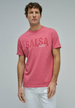 Hombre con cabello rizado vistiendo una camiseta rosa de SALSA JEANS y pantalones blancos, posando sobre un fondo liso de color gris claro.