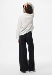 Pull blanc texturé à manches longues, associé à un jean noir taille haute à jambes larges. Le mannequin pose avec une main dans les cheveux.