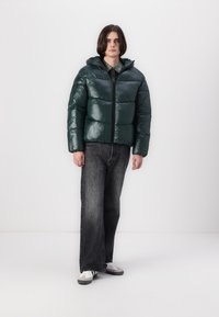 Chaqueta acolchada verde con acabado brillante, cierre de cremallera y capucha. Llevada con jeans de pierna ancha negros y zapatillas blancas, sobre un fondo simple.