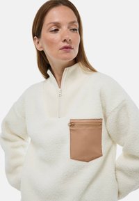 Pull-over en polaire blanche avec un col montant à côtes, une fermeture éclair sur le devant à quart et une poche zippée beige sur le côté gauche. Tissu doux et texturé.