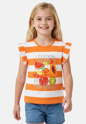 Jeune fille souriante, portant un débardeur rayé orange et blanc avec des illustrations colorées de fruits et un short en jean.