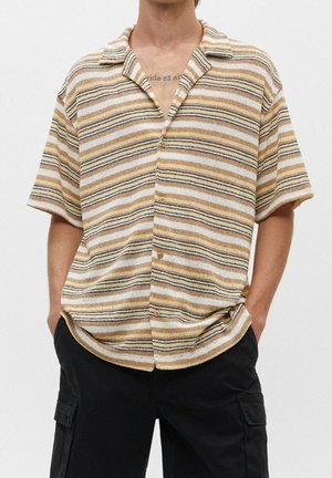 Homme portant une chemise à manches courtes à rayures beige, blanche et noire, boutonnée, avec un pantalon cargo noir, debout les mains dans les poches.