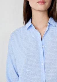 Camicia azzurro chiaro con bottoni, caratterizzata da un motivo a griglia testurizzato, collettino e bottoni bianchi. Il tessuto sembra liscio e leggero.
