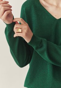 Zaļš, strukturēts adījuma pulovers ar V veida izgriezumu un garām piedurknēm. Roku rotā zelta gredzens ar gludu, spīdīgu virsmu.