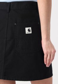 Čierna džínová sukňa s vreckom na zadnej strane s bielym logom Carhartt, zapínanie na gombík a viditeľnými švami.