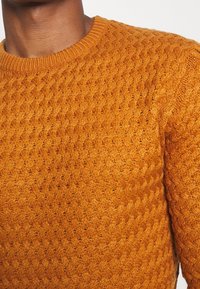 Gros plan sur une personne portant un pull moutarde texturé en maille avec un col rond.