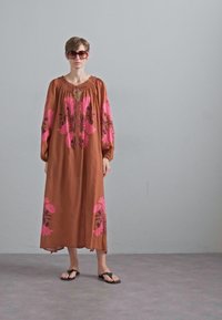 Lang bruine jurk met roze bloemenborduursel, poffende mouwen en een V-hals. Gemaakt met zwarte teenslippers en oversized zonnebril.