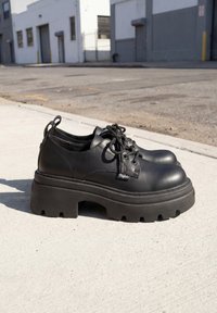 Schwarze Leder-Chunky-Plateau-Schnürschuhe auf Betonweg mit Industriegebäuden im Hintergrund.