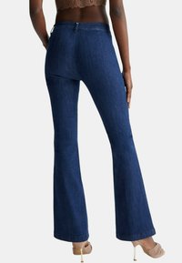 Jeans a zampa d'elefante in denim blu scuro con vita media, dettagli di cucitura sottili e una texture morbida, abbinati a scarpe con tacco trasparenti.
